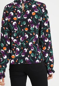 Langærmet blomsterbluse med en sort baggrund og lilla, orange og hvide blomster. Flæsekrave og manchetter med en nøglehulbagdetalje.