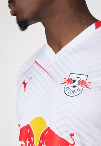 Bijela nogometna majica s crvenim naglascima, s v-izrezom, teksturiranim uzorkom i logotipom RB Leipziga na grudima.