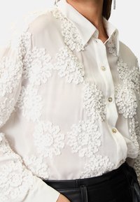 Blouse blanche en tissu transparent avec des appliqués floraux texturés, un col classique et une fermeture à boutons. Les détails incluent des boutons semblables à des perles.