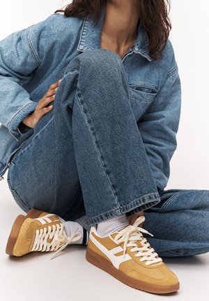 Personne portant des baskets marron clair avec des accents blancs, des chaussettes blanches, un jean bleu en denim et une veste en denim bleue, assise en tailleur sur un sol blanc.