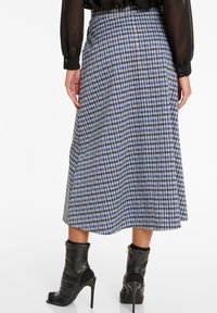 Midikleid aus blauem und schwarzem Hahnentrittmuster in fließender Silhouette, mit Taillennaht und seitlichen Falten. Tragen mit schwarzen Absatzstiefeln.