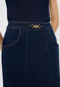 Tummansinisestä denim-hameesta, jossa on kullanvärinen solki vyötäröllä. Hameessa on kaksi kaarevaa taskua ja näkyviä ompeluviivoja.