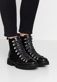 Czarne botki za kostkę z mieszanką gładkiej skóry i zamszu, z metalowymi elementami w kolorze srebrnym, grubymi gumowymi podeszwami oraz kontrastującymi sznurowadłami.