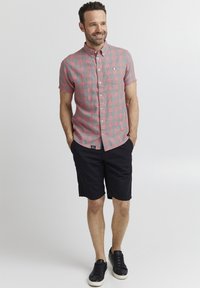 Camicia a maniche corte a quadri rossa e grigia, pantaloni corti neri e sneakers nere. La camicia ha un collerino a bottoni e una tasca sul petto.