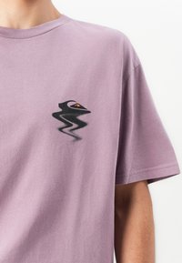 Lilla lühikese varrukaga T-särk, millel on vasakul rinnal moonutatud must ja oranž abstraktne muster, kantud inimese poolt.