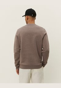 Brauner Pullover mit Rundhalsausschnitt, gerippten Bündchen und Saum. Glatte Textur mit lockerer Passform, getragen zu hellen Hosen und einer schwarzen Mütze.