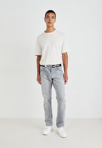 Lichtgrijze jeans met een relaxte pasvorm, gecombineerd met een eenvoudig wit T-shirt. Een zwarte riem met vetgedrukte witte tekst en witte sneakers maken de outfit af.