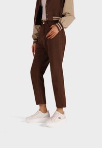 Jeans marroni a vita alta con una vestibilità dritta. Abbinati a sneakers bianche con zeppa e dettagli metallici e accenti rosa.