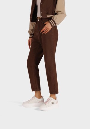 Jeans marroni a vita alta con una vestibilità dritta. Abbinati a sneakers bianche con zeppa e dettagli metallici e accenti rosa.