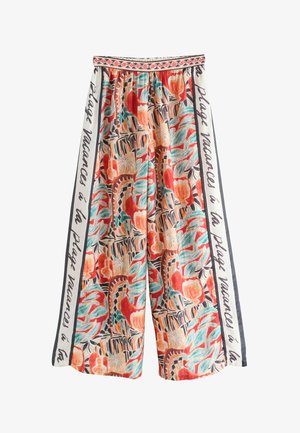 Pantalon à jambes larges avec imprimé tropical abstrait coloré en rouge, bleu sarcelle et beige, avec des panneaux latéraux blancs avec texte en français et une ceinture à motifs.