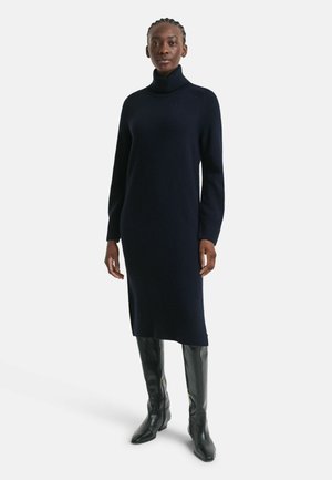 Navy turtleneck dress made of knit material; features long sleeves, a fitted silhouette, and side slits, paired with tall black boots.