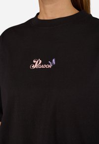 Black cotton T-shirt featuring a pink cursive "Pegador" logo and a small purple butterfly graphic on the left chest area.