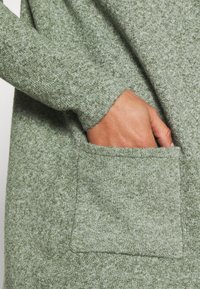 Zelená pletená tkanina s jemnou texturou má přední kapsu a dlouhé rukávy. Barva je tlumená, melírovaná zelená.