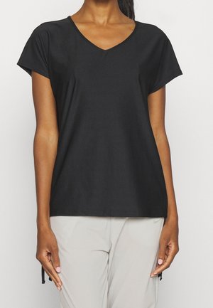 Femme portant un t-shirt noir uni à col en V et manches courtes et un pantalon clair, debout devant un fond uni.