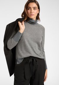 Grauer Rollkragenpullover mit glatter Textur und geripptem Kragen, kombiniert mit schwarzen Hosen mit gebundenem Bund und einer Jacke mit Streifenmuster.