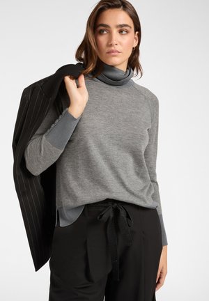 Grauer Rollkragenpullover mit glatter Textur und geripptem Kragen, kombiniert mit schwarzen Hosen mit gebundenem Bund und einer Jacke mit Streifenmuster.