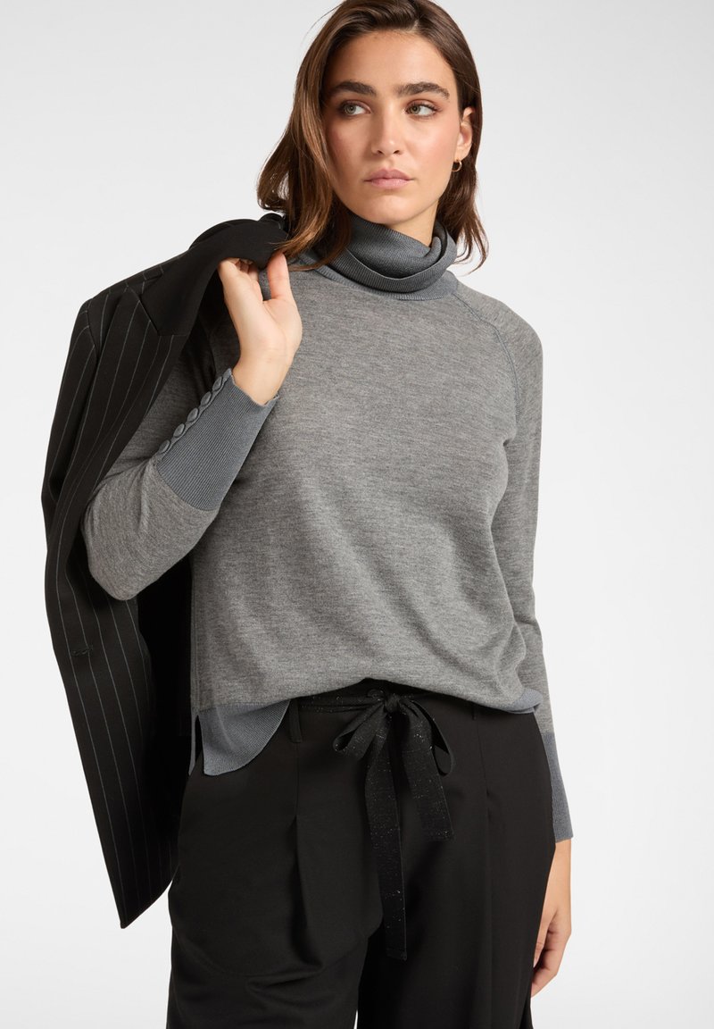 Grauer Rollkragenpullover mit glatter Textur und geripptem Kragen, kombiniert mit schwarzen Hosen mit gebundenem Bund und einer Jacke mit Streifenmuster.