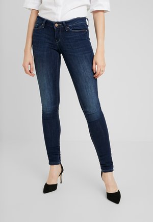 Femme portant un jean skinny bleu foncé et des talons hauts noirs, chemise blanche coupée visible de la taille vers le bas sur fond gris.