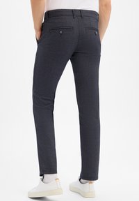 Man in een donkergrijze slim-fit broek met vastgemaakt achterzakken, witte sokken en witte sneakers, met zijn handen in de zakken, van achteren gezien.