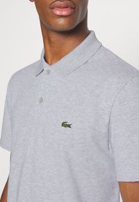 Szara koszulka polo z bawełny, z kołnierzykiem, dwoma guzikami i haftowanym zielonym logo krokodyla na lewej piersi.