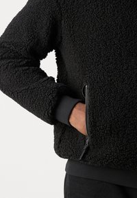 Giacca in pile nera con una superficie morbida e testurizzata; presenta una tasca con zip e polsini a coste per una vestibilità aderente.