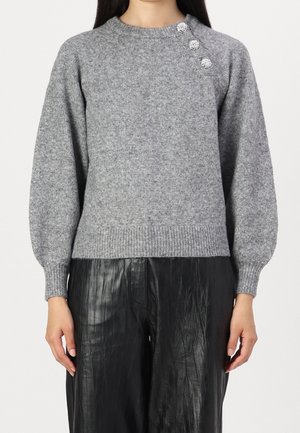 Žena v šedém pleteném svetru s třemi ozdobnými knoflíky na rameni a černých texturovaných kalhotách, na jednobarevném pozadí.