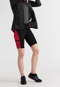 Persona vistiendo pantalones cortos de ciclismo negros y rojos, camiseta gris, chaqueta negra y zapatillas de correr negras, de pie contra un fondo claro y sencillo.