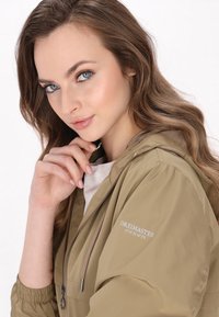 Giacca a vento khaki chiaro con chiusura a zip, cappuccio regolabile con cordino e polsini elastici. Presenta un logo sulla manica in caratteri grigi.
