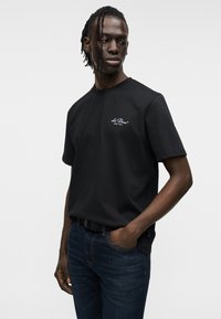 Μαύρο βαμβακερό T-shirt με στρογγυλή λαιμόκοψη και κοντά μανίκια. Φέρει ένα μικρό μπλε λογότυπο στο στήθος. Συνδυάζεται με σκούρο τζιν παντελόνι.