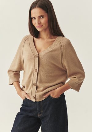 Mujer con cabello largo y castaño vistiendo un cárdigan beige abotonado y jeans oscuros de mezclilla con las manos en los bolsillos, sonriendo ligeramente.