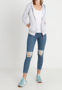 Sudadera con cremallera de color gris claro, con un bolsillo frontal, combinada con unos jeans ajustados de color azul con rotos en las rodillas, y zapatillas de color verde menta con acentos blancos.