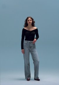 Must pikkade varrukatega topp keeratud esiosaga, millele on kombineeritud helesinised kõrge vöökohaga teksad. Modell seisab ühtlase helesinise tausta ees.