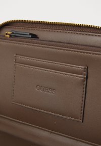 Brown leather wallet with a zip closure, featuring a card slot and embossed "GUESS" logo, detailed with gold hardware and stitching.
