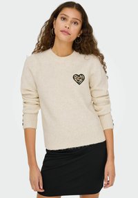 Knit džemperis krēmkrāsā ar leoparda raksta sirdspoguļa plāksteri uz krūtīm. Iezīmējas ar ribotu tekstūru un pogu akcentiem uz piedurknēm. Apvienots ar melnu svārku.