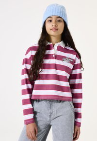 Gestreiftes langärmliges Polo in Burgunderrot, Pink und Hellblau, mit einem weißen Kragen und dem Logo "Trouble Maker" auf der Brust. Dazu werden graue Jeans getragen.