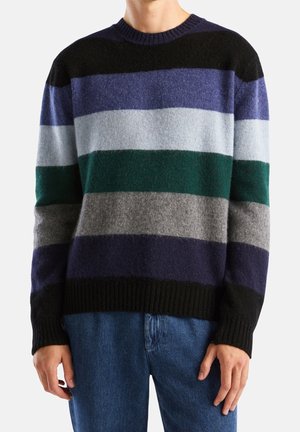 Mężczyzna ubrany w wełniany sweter z poziomymi paskami w kolorach czarnym, fioletowym, jasnoniebieskim, zielonym, szarym i granatowym, w połączeniu z niebieskimi dżinsami.