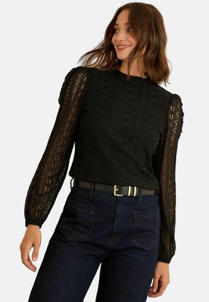 Schwarze Spitzenbluse mit langen durchsichtigen Ärmeln und strukturiertem Oberteil. Sie hat einen runden Hals und wird mit dunklen Jeans kombiniert, die durch einen Gürtel akzentuiert werden.