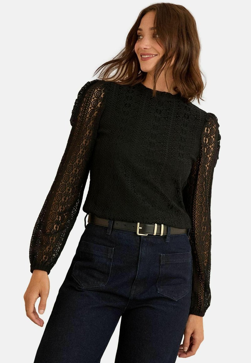 Fekete csipkeblúz hosszú átlátszó ujjal és texturált testtel. Kerek nyaka van, és sötét farmernadrággal párosítják, övvel kiemelve.