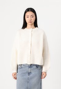 Krémová pletená cardigan s ribovanou textúrou, s okrúhlym výstrihom a šiestimi veľkými gombíkmi na prednej strane. Dlhé, mierne nariasené rukávy.