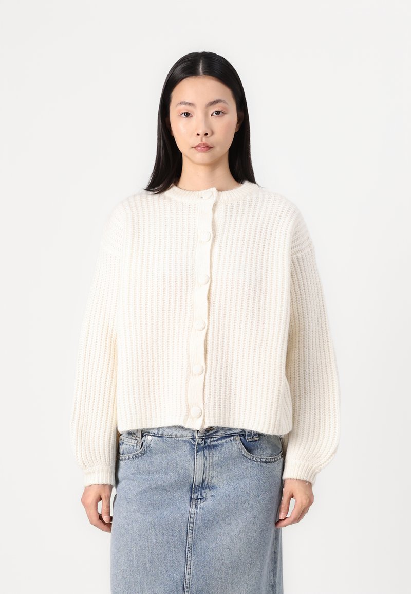 Krémová pletená cardigan s ribovanou textúrou, s okrúhlym výstrihom a šiestimi ve�ľkými gombíkmi na prednej strane. Dlhé, mierne nariasené rukávy.