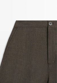 Bukser i mørkebrunt linnedstof med frontlomme og knaplukning i taljebåndet, vist i nærbillede.