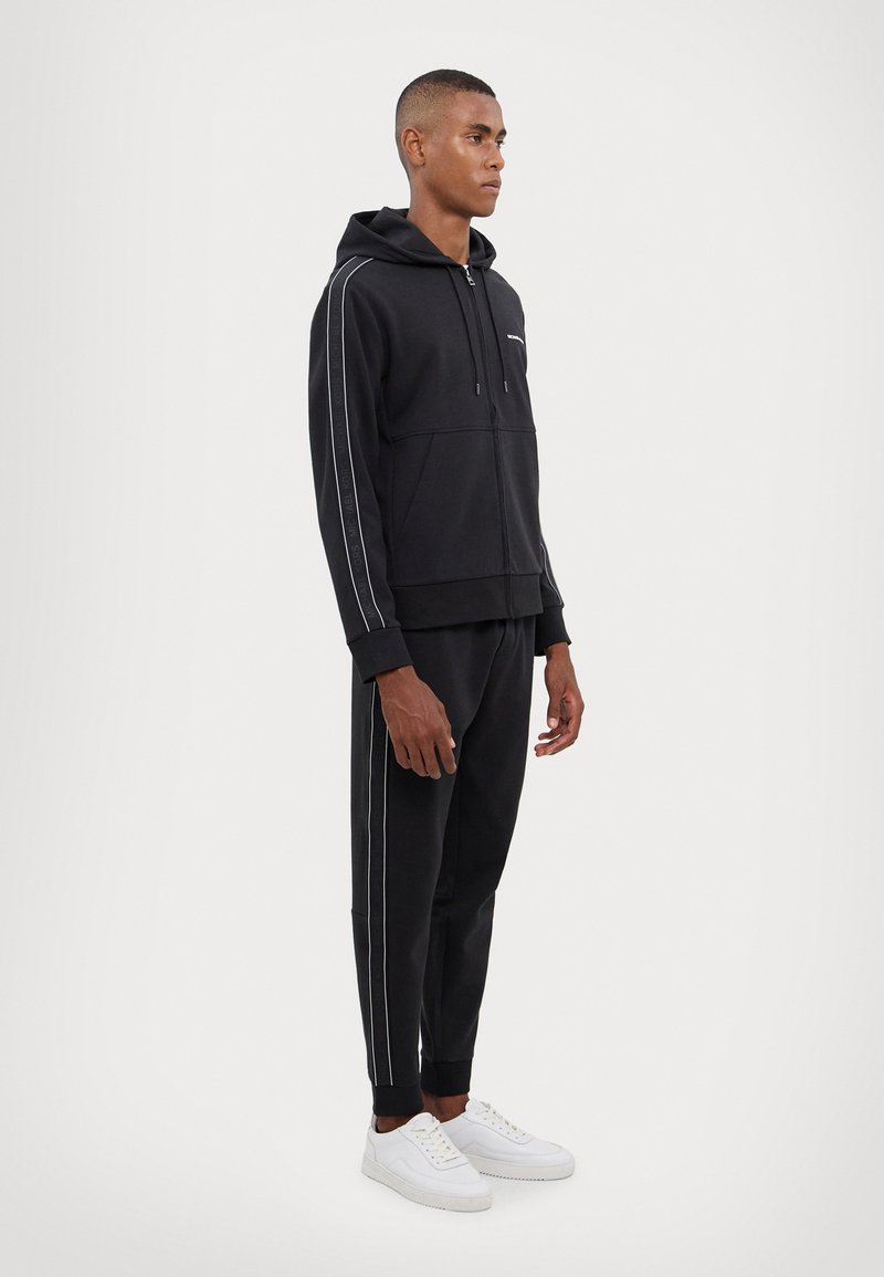 Black hoodie and sweatpants set made of cotton blend, featuring white side stripes, kangaroo pocket, and ribbed cuffs. White sneakers complete the look.