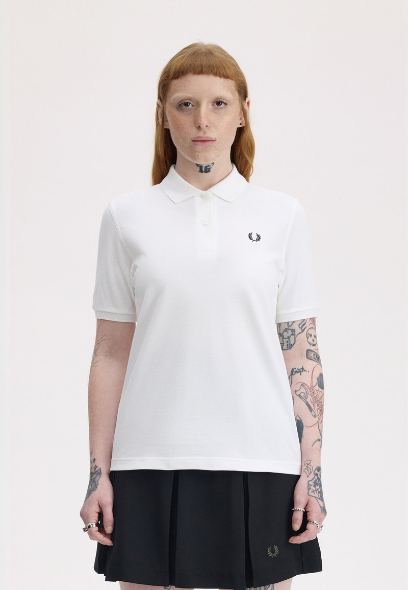 Personne aux cheveux roux avec des tatouages sur le cou et les bras, portant un polo blanc et une jupe plissée noire, debout devant un fond clair uni.