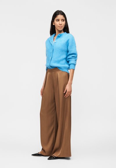 Γυναίκα που φοράει γαλάζια ζακέτα με υφή, καφέ παντελόνια φαρδιάς γραμμής και μαύρα μυτερά φλατ παπούτσια, στέκεται μπροστά σε απλο φόντο.