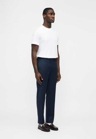 Άνδρας που στέκεται, φορώντας λευκό κοντομάνικο μπλουζάκι, ναυτικά ραμμένο παντελόνι, λευκές κάλτσες και σκούρα καφέ δερμάτινα μοκασίνια, σε απλό φόντο.