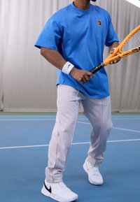Blue cotton t-shirt with a logo patch, light gray athletic pants with side stripes, and white sneakers with black accents. Tennis racket held.