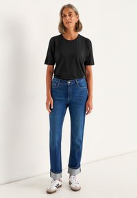 Sort kortærmet t-shirt, blå lige ben jeans med opfoldede nederste kanter, hvide sneakers med sorte og grå detaljer, båret af en model i en neutral indstilling.