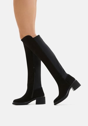 Schwarze Überknie-Stiefel aus einer Kombination von Wildleder und elastischem Material, mit einem seitlichen Reißverschluss und einem dicken Absatz. Durchgehend glatte Textur.