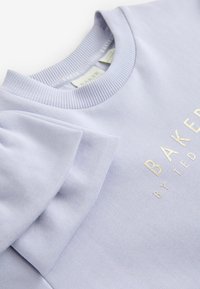 Šviesiai violetinė palaidinė, pagaminta iš minkštos medžiagos. Turi raštuotą kaklą ir sluoksniuotas pūstas rankoves. Aukso spalvos užrašas sako "BAKER BY TED."