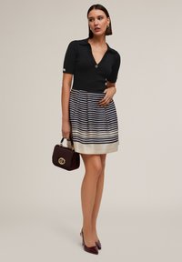 A black ribbed top with a V-neck and short sleeves paired with a striped pleated skirt in navy and cream, holding a burgundy handbag.